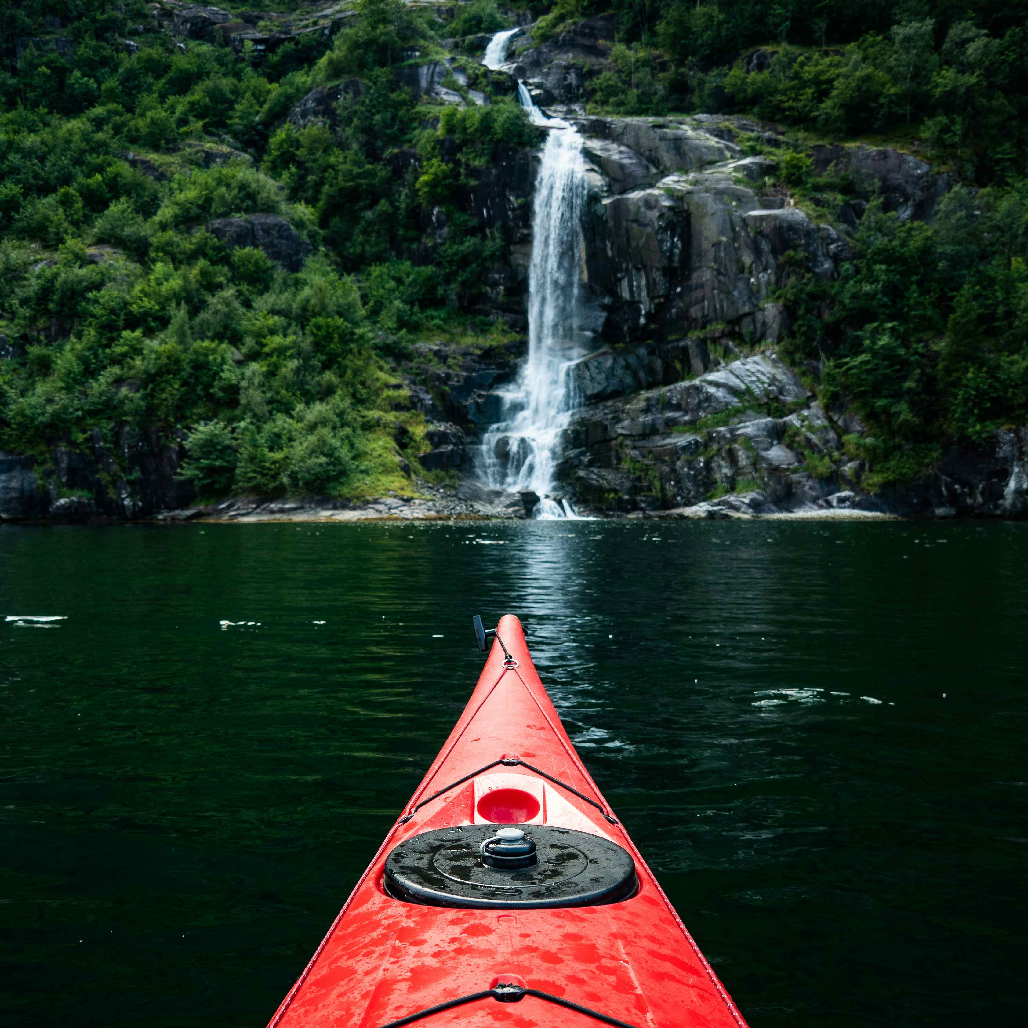 The tip of a red kayak in the blue fjord. A waterfall is seen in the distance from the mountainside.